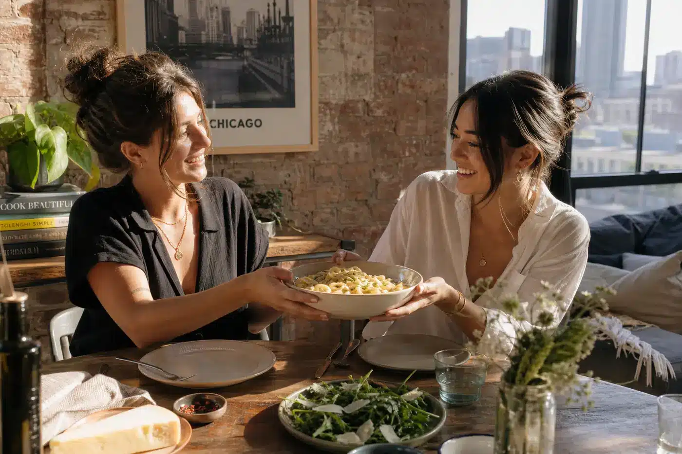 Dinner Ideas for Friend Gathering: Stress-Free Hosting Tips Two friends sharing a bowl of fresh pasta at a wooden dining table in a sunlit Chicago loft